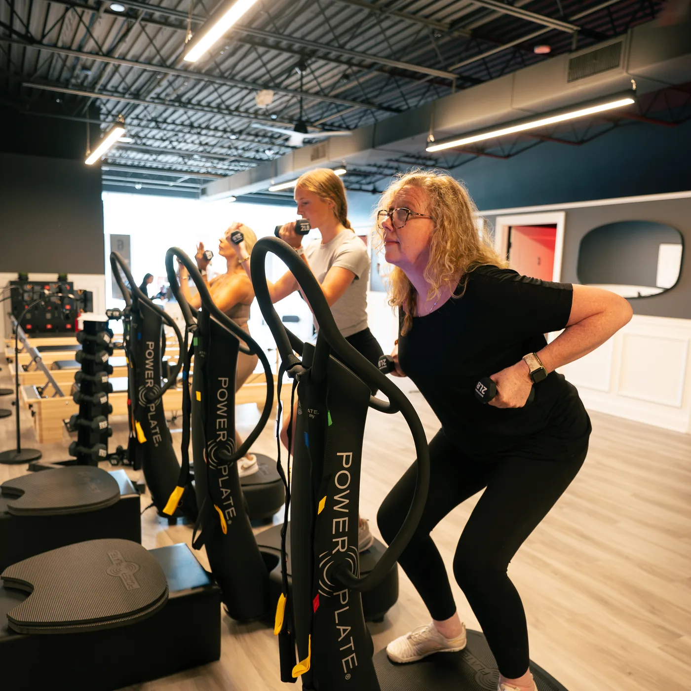 Two women standing on whole-body vibration platforms on an open studio floor